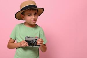 knap jongen in zomer hoed Holding een wijnoogst retro camera, looks Bij de camera poseren tegen roze achtergrond. toerisme concepten foto