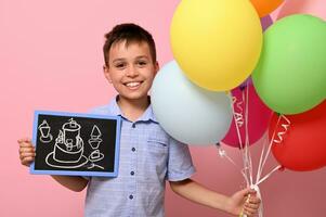 schattig jongen houdt veelkleurig kleurrijk ballonnen in een hand- en een schoolbord met getrokken verjaardag taart in de ander. geïsoleerd over- roze achtergrond met kopiëren ruimte foto