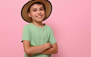 gelukkig knap jongen in zomer rietje hoed glimlacht met toothy glimlach , staand met gekruiste armen Aan roze achtergrond terwijl poseren naar camera. kinderen emoties. foto