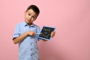 glimlachen school- jongen Holding een schoolbord met belettering terug naar school- en richten met een potlood Aan het, poseren over- roze achtergrond met ruimte voor tekst foto