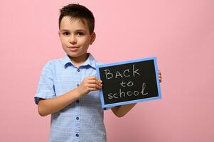 aanbiddelijk schooljongen Holding een schoolbord met krijt belettering ,rug naar school, geïsoleerd over- roze achtergrond met ruimte voor tekst foto