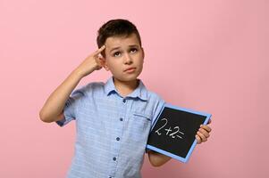 een schooljongen met een schoolbord in zijn hand- houdt een vinger naar zijn tempel en met een attent kijken lost op een rekenkundig probleem, staand Aan een roze achtergrond met een plaats voor tekst. terug naar school. foto