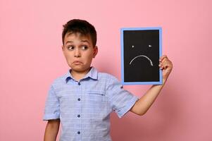 geïsoleerd portret Aan roze achtergrond van een verdrietig schooljongen Holding in de buurt zijn gezicht een bord met getrokken verdrietig emotie. ruimte voor tekst foto