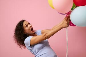 studio schot voor reclame van een verrast vrouw bereiken voor kleurrijk lucht ballonnen, staand tegen roze achtergrond met kopiëren ruimte. de concept van viering, vakantie, verjaardag en geschenken. foto