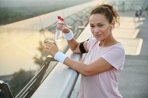 sportvrouw in een roze t-shirt en hoofdtelefoons houdt een fles van water en bewondert de mooi keer bekeken van de rivier- en natuur, staand Aan de stad brug, resting na een ochtend- joggen in de vers lucht foto