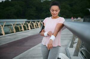 aantrekkelijk spaans vrouw in hoofdtelefoons en een roze t-shirt staat Aan een stad brug en draagt badstof tennis polsbandjes voordat ochtend- joggen en cardio opleiding. mooi vrouw atleet werken uit buitenshuis foto