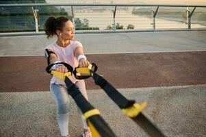 voorkant visie van Afrikaanse Amerikaans sportvrouw aan het doen squats met suspensie riemen, beoefenen lichaam gewicht fysiek opdrachten buitenshuis Aan mooi vroeg zomer dag. sport, fitheid, gewicht verlies concept foto