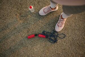 hoog hoek visie van vrouw atleet poten vervelend roze sportschoenen en staand Aan een asfalt loopband De volgende naar een aan het liegen naar beneden overslaan touw en water fles foto