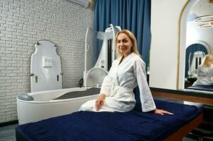 aantrekkelijk blond vrouw in wit badjas zit Aan massage tafel en wacht voor schoonheid behandelingen in spa met modern multifunctioneel spa capsule foto