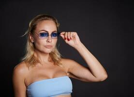 zelfverzekerd portret van een charmant midden- oud Europese vrouw met golvend blond haar- vervelend een blauw top en blauw zonnebril en op zoek Bij camera geïsoleerd over- zwart achtergrond met kopiëren advertentie ruimte. foto