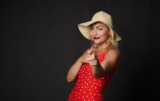 aantrekkelijk blond Kaukasisch vrouw in rood kleding met wit polka dots en rietje zomer hoed glimlacht mooi wit toothy glimlach, richten omhoog vinger Bij kopiëren ruimte Aan zwart achtergrond foto