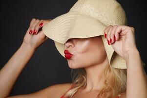 detailopname mooi Kaukasisch vrouw aan het bedekken haar ogen met een rietje zomer hoed en Bezig met verzenden kus, geïsoleerd over- zwart achtergrond met kopiëren ruimte. hoofdschot portret van een mooi dame met rood lippenstift foto