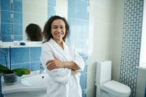 volwassen Afrikaanse Amerikaans vrouw in wit wafel badjas leunend Aan wastafel in badkamer en glimlachen Bij camera met gekruiste armen foto