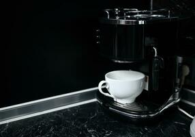 zwart elektrisch koffie machine met wit keramisch kop Bij keuken aanrecht foto