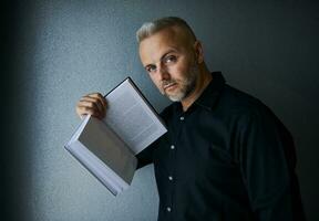 portret van indrukwekkend imposant knap elegant midden- oud Kaukasisch Mens, geslaagd zakenman, ondernemer Holding een boek, op zoek Bij camera poseren tegen grijs muur achtergrond met kopiëren ruimte foto