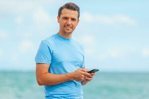 jong Mens Holding mobiel telefoon in zijn hand- terwijl wandelen Aan de strand foto