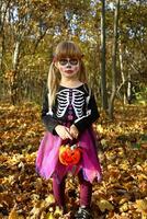 weinig meisje in maskerade kostuum voor dag van dood, dia de los muertos, met suiker schedel verzinnen. halloween skelet zwart en paars jurk. Holding oranje pompoen plastic emmer met snoepjes. verticaal. foto