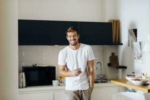 portret van gelukkig jong Mens Holding cel telefoon in keuken foto