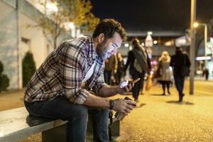 uk, Londen, glimlachen Mens zittend Aan een bank en op zoek Bij zijn telefoon door nacht foto