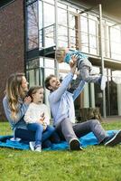 gelukkig familie Aan deken in tuin in voorkant van hun huis met vader lichtgevend omhoog zoon foto