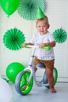 een weinig jongen met een pet Aan zijn hoofd zit achter een balans fiets tegen de achtergrond van groen ballonnen. verjaardag voor kinderen. vieren st. Patrick dag. geschenk fiets voor kind foto