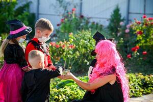 kinderen truc of traktatie in halloween kostuum en medisch masker. een weinig jongen, meisje en baby in pakken gedurende de coronavirus pandemisch te ontvangen snoep van een vrouw. foto