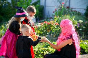 kinderen truc of traktatie in halloween kostuum en medisch masker. een weinig jongen, meisje en baby in pakken gedurende de coronavirus pandemisch te ontvangen snoep van een vrouw . foto