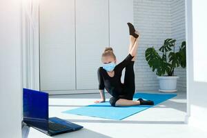 fit weinig meisje in medisch masker aan het doen yoga en aan het kijken online lessen Aan laptop, oefenen in de kamer. foto