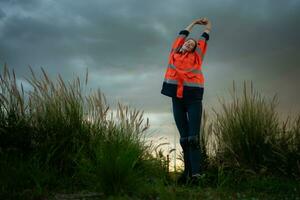 jong vrouw in ingenieur uniform en hoog zichtbaarheid met verheven armen staand Aan met gras begroeid veld- Bij zonsondergang, de concept van kom tot rust tijd na werk foto