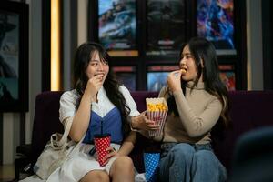 twee jong Aziatisch Dames aan het eten popcorn en aan het wachten voor aan het kijken film Bij voorkant van bioscoop foto