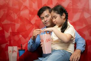 vader en dochter aan het eten popcorn en aan het wachten voor aan het kijken film Aan rood achtergrond. familie vermaak concept. foto