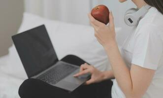 doeltreffend jong millennial meisje zittend Aan een bed in de ochtend, toepassingen laptop computer en eet croissants en drankjes koffie voor ontbijt foto