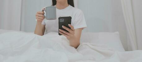 doeltreffend jong millennial meisje zittend Aan een bed in de ochtend, toepassingen laptop computer en eet croissants en drankjes koffie voor ontbijt foto