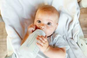 schattig weinig pasgeboren meisje drinken melk van fles en op zoek Bij camera Aan wit achtergrond. zuigeling baby zuigen aan het eten melk voeding aan het liegen naar beneden Aan wieg bed Bij huis. moederschap gelukkig kind concept. foto