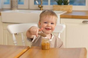 gelukkig familie Bij huis. baby jongen voeden zichzelf in keuken. weinig jongen met rommelig grappig gezicht eet gezond voedsel. kind leert eten door zichzelf Holding lepel. zelf voeden. foto