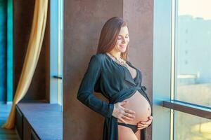 zwangerschap vrouw staand in de buurt venster Bij huis en glimlacht foto