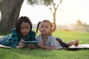 twee jong meisjes aan het liegen Aan de gazon op zoek Bij hun telefoons, zomer, gouden uur, zonsondergang. sstkhuis foto