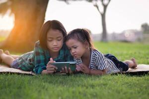 twee jong meisjes aan het liegen Aan de gazon op zoek Bij hun telefoons, zomer, gouden uur, zonsondergang. sstkhuis foto
