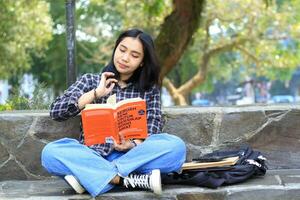 gelukkig bedachtzaam jong Aziatisch vrouw college leerling lezing een boek in de park, onderwijs concept foto