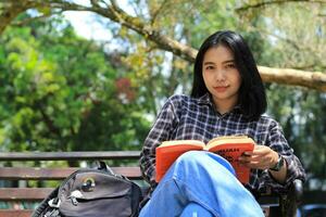 gelukkig jong Aziatisch vrouw college leerling met glimlachen gezicht genieten lezen een boek voor tentamen in de park foto