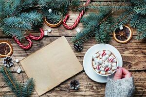 een kop van heet chocola met heemst in de handen van een Mens in een trui. Kerstmis boom en decoraties, riet karamel en sinaasappels noten houten achtergrond kopiëren ruimte. nieuw jaar geschenk. foto