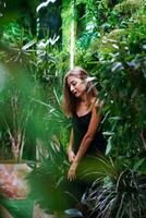 meisje op zoek naar beneden bedachtzaam. regenwoud. jong vrouw vervelend zwart jurk Aan dun riemen staat tussen groen planten binnen regeling. jong mooi vrouw model- poses tussen groen tropisch planten. foto