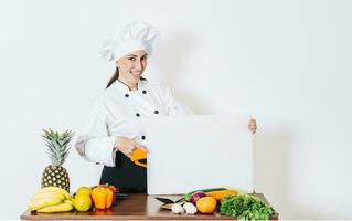 chef vrouw met groenten Bij tafel Holding blanco menu. meisje chef in de keuken tonen een blanco bord, mooi vrouw chef met tafel van groenten Holding een blanco bord foto