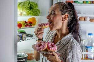 hongerig brunette in pyjama geniet zoet donuts laat Bij nacht door de Open koelkast foto