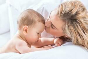 moeder en baby jongen zoon spelen Aan een wit bed. moeders tederheid en kusjes van een kleuter kind foto