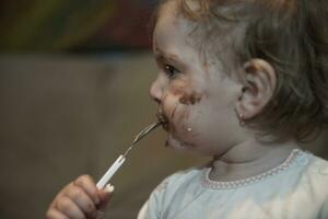 baby meisje aan het eten haar chocola woestijn met een lepel en maken een knoeien foto