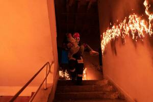 dapper brandweerman daalt af trap van een brandend gebouw en houdt opgeslagen meisje in zijn armen. Open brand en een brandweerman in de achtergrond. foto