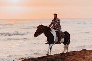 een modern Mens in zomer kleren geniet rijden een paard Aan een mooi zanderig strand Bij zonsondergang. selectief focus foto