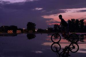 eenzaam kinderen silhouet Aan fiets, jongen rijden fiets Aan reflecterende water. achtergrond mooi zonsondergang. foto