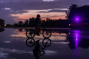 eenzaam kinderen silhouet Aan fiets, jongen rijden fiets Aan reflecterende water. achtergrond mooi zonsondergang. foto